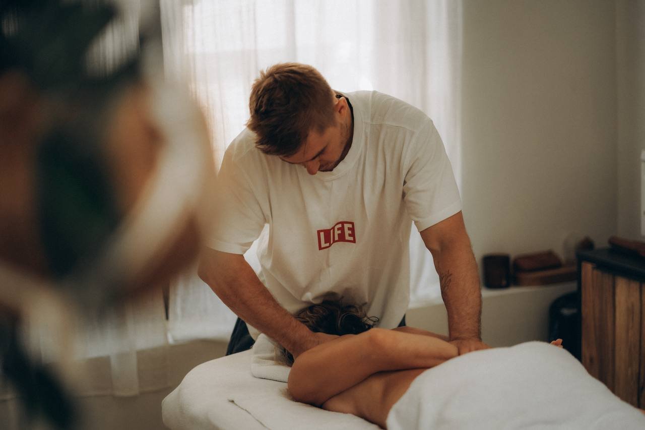 A man performing a massage on a woman lying face down on a massage table in a room with natural light.